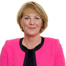 A lady facing the camera in a black top and bright pink jacket, smiling slighlty.